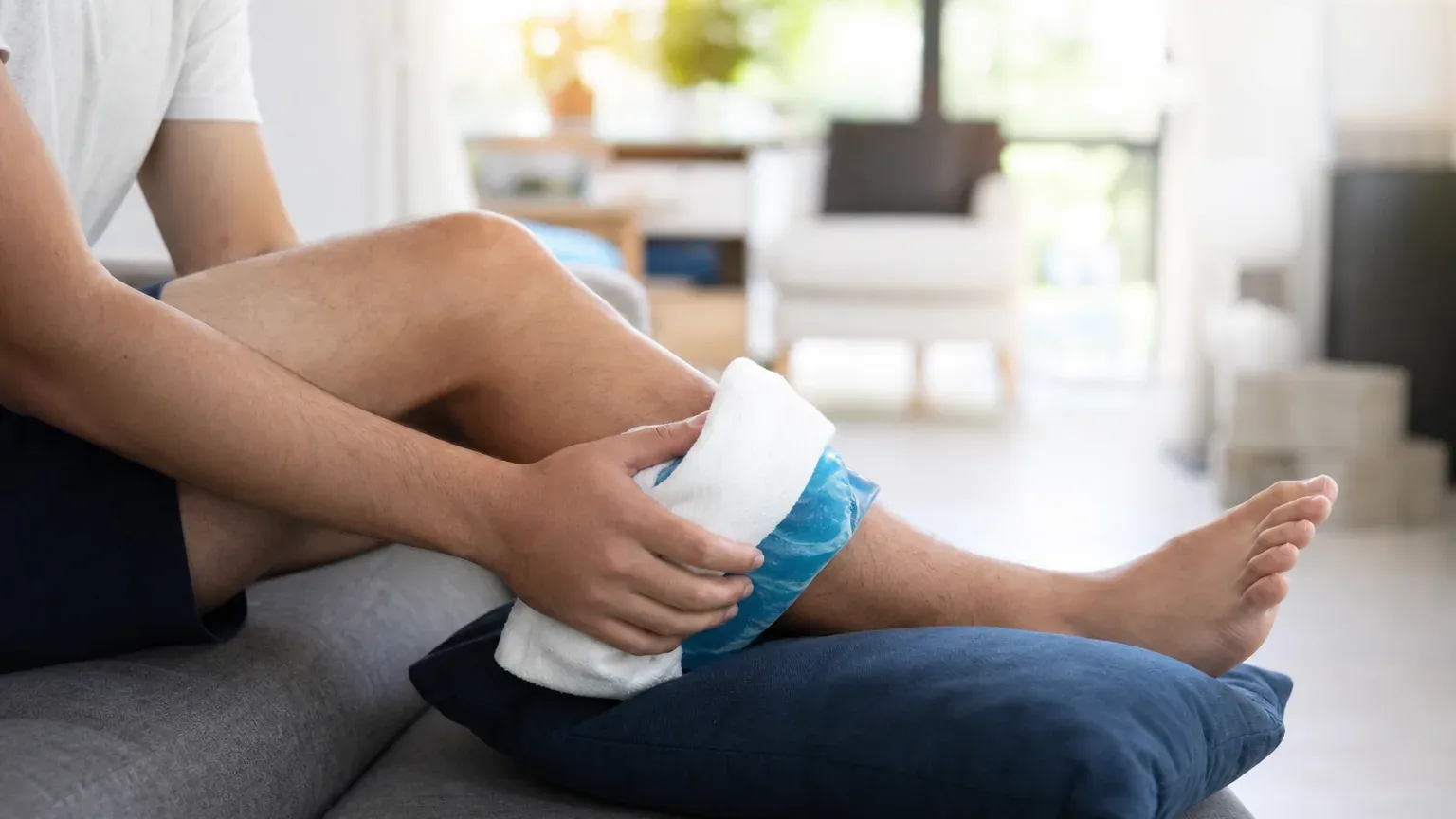 Personne appliquant une poche de froid sur sa cheville, jambe surélevée sur un coussin