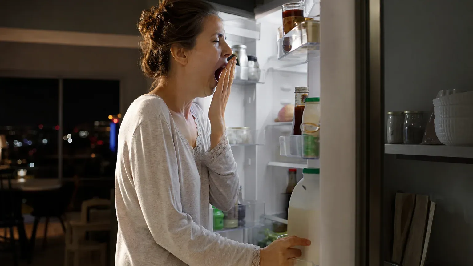 Personne bâillant devant le réfrigérateur ouvert tard le soir dans une cuisine faiblement éclairée